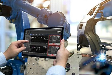 Someone holding a tablet displaying welding data; a car body is welded in the background. 