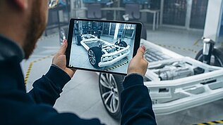 Person with tablet in hand, in the background the basic structure of an electric vehicle. 